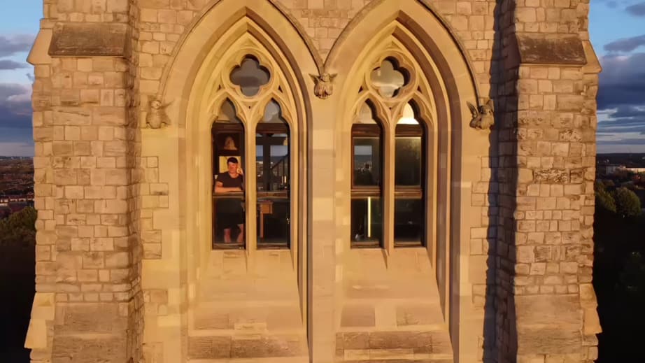 Die Turmfassade im Abendlicht — zwei hohe gotische Bögen mit Vierpassmaßwerk und geschnitzten Engelskonsolen, dahinter Delehag klein im erleuchteten Fenster