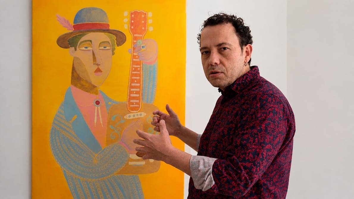Felix Zilinskas neben einer hochformatigen gelben Leinwand mit einer behuteten, blau gekleideten Figur, die eine orangefarbene, wie eine Streichholzschachtel gebaute Gitarre hält, die Hände mitten in der Erklärung erhoben