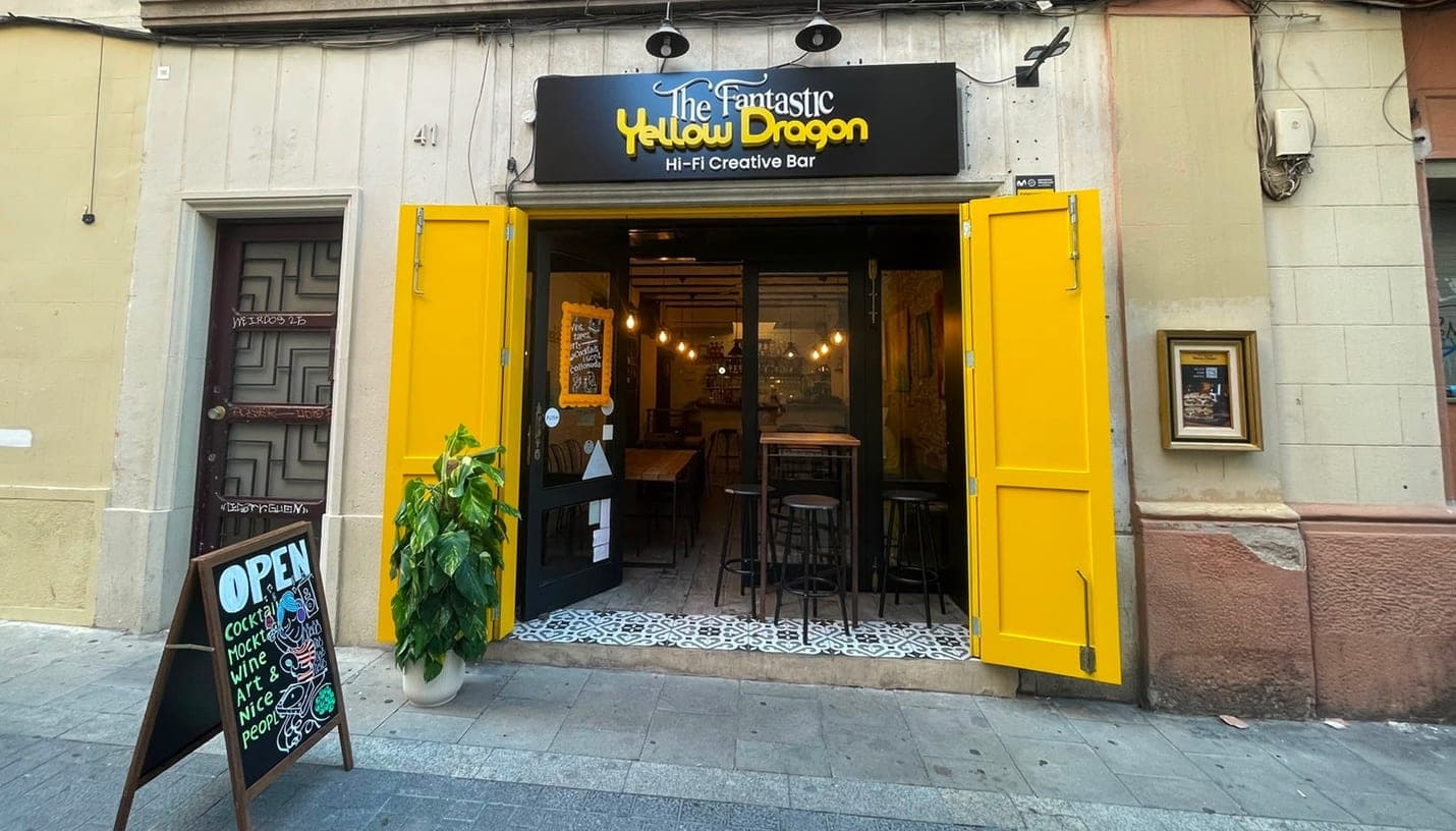 Straßenansicht von The Fantastic Yellow Dragon, der Hi-Fi-Kreativbar in Gràcia — weit geöffnete gelbe Fensterläden und eine Tafel auf dem Gehweg mit der Aufschrift Cocktails, Mocktails, Wine, Art & Nice people